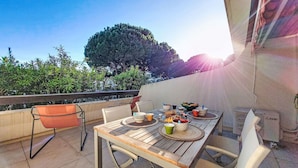 Un área de comedor al aire libre con una mesa preparada para dos personas, rodeada de sillas, en una terraza con vistas a un árbol grande y un cielo despejado.