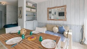 Un comedor con una mesa de madera dispuesta para cuatro personas, rodeada de sillas blancas. Un espejo grande cuelga en la pared, y hay un mueble empotrado con cajas de almacenamiento.