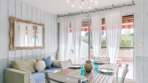 Una habitación con una mesa de madera dispuesta para dos personas, un sofá con cojines, un espejo en la pared y un balcón con cortinas.