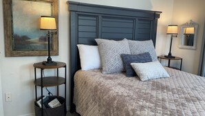 Une chambre à coucher avec un grand lit, une tête de lit, des tables de chevet avec des lampes, un tableau accroché au mur et une petite table d’appoint avec un panier.