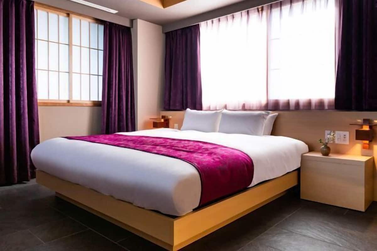 A modern hotel room with a large bed, a nightstand, a bedside table, and a window with curtains.
