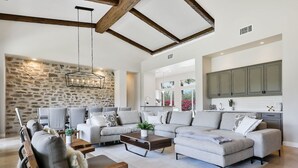A spacious living room with a stone wall, a wooden ceiling, and a modern kitchen in the background. - Casa Del Sol - Private Courtyard w/ Pool & Incredible views of PGA West. #241306 6br (La Quinta)