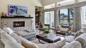 A spacious living room with a stone fireplace, comfortable seating, and a large TV displaying a golf game. - Casa Del Sol - Private Courtyard w/ Pool & Incredible views of PGA West. #241306 6br (La Quinta)