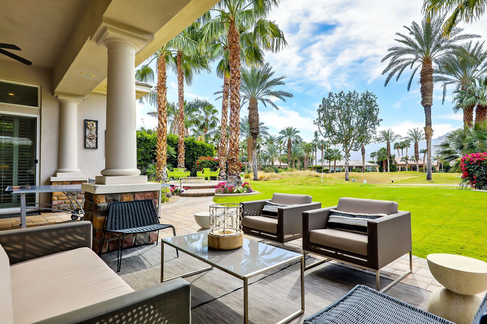 An outdoor patio with seating, a stone fireplace, and a view of a well-maintained lawn and palm trees.