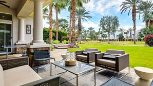 An outdoor patio with seating, a stone fireplace, and a view of a well-maintained lawn and palm trees. - Casa Del Sol - Private Courtyard w/ Pool & Incredible views of PGA West. #241306 6br (La Quinta)