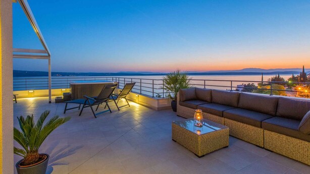 A rooftop terrace with a sofa, chairs, and a table, overlooking a body of water and cityscape at dusk. - Holiday apartment near the beach with shared hot tub (Dramalj)