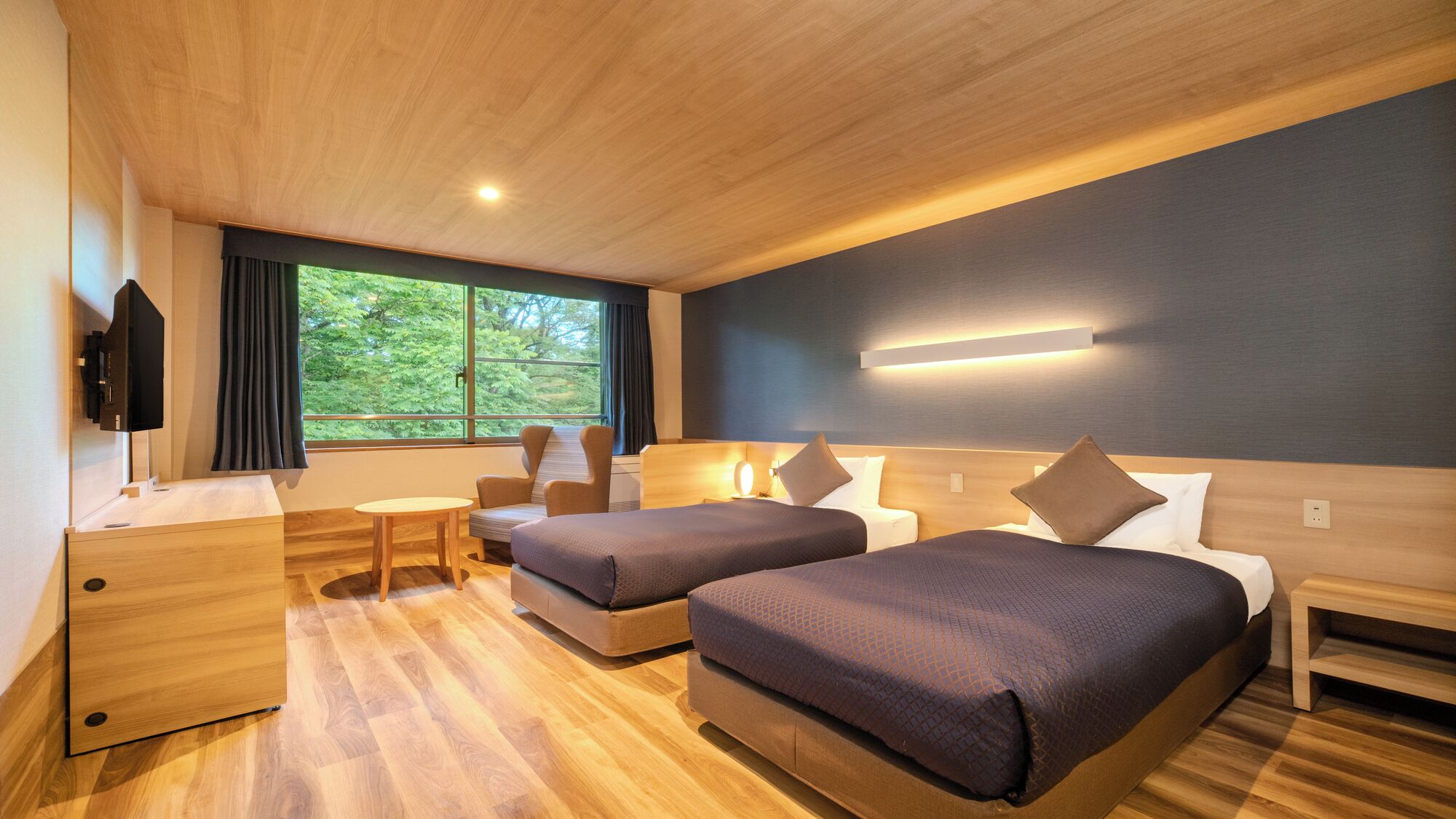 Hotelkamer met twee bedden, een bureau, een stoel, een televisie en een groot raam met uitzicht op groen.