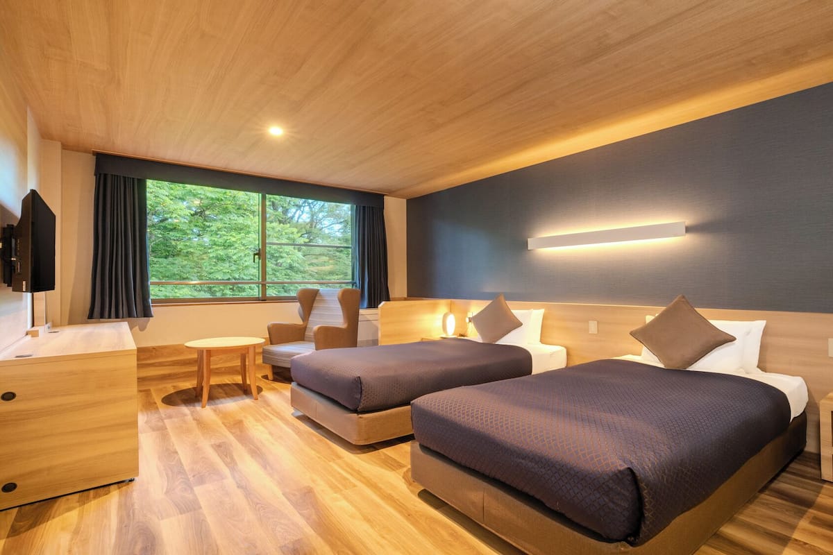 Habitación de hotel con dos camas, un escritorio, una silla, un televisor y un ventanal con vistas a zonas verdes.