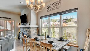 A dining area with a stone fireplace, wooden chairs, and a large window offering a view of houses.