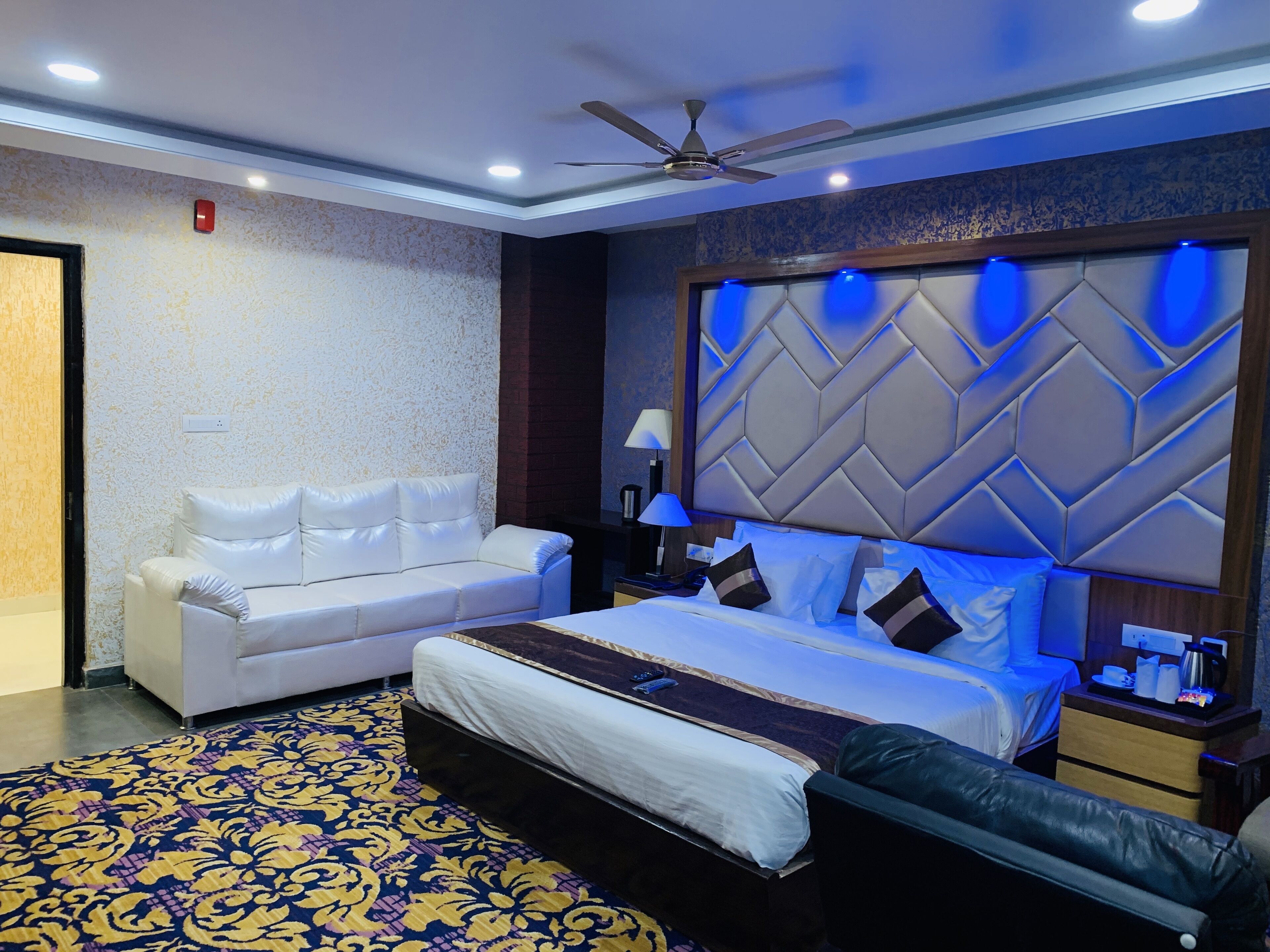 Ein modernes Hotelzimmer mit einem großen Bett, einem weißen Sofa, einem gemusterten Teppich und einem Deckenventilator.
