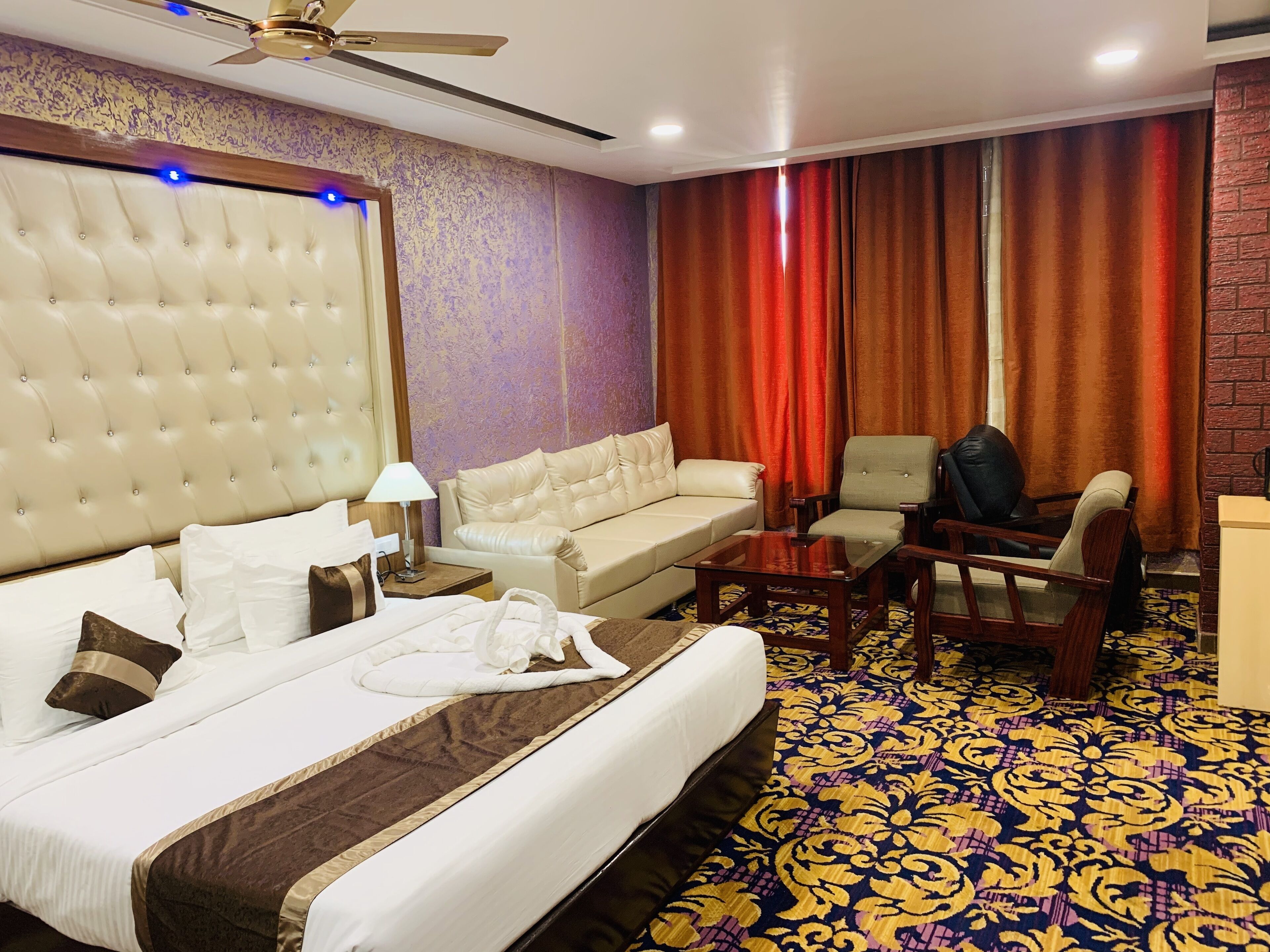 A hotel room with a large bed, a sofa, a chair, a small table, and a ceiling fan.