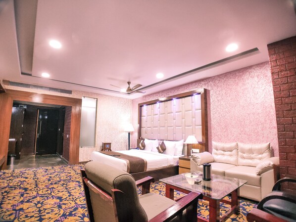 Habitación de hotel con cama, sofá, mesa de centro y una silla. Se observa una puerta que conduce a otra habitación y una alfombra estampada.