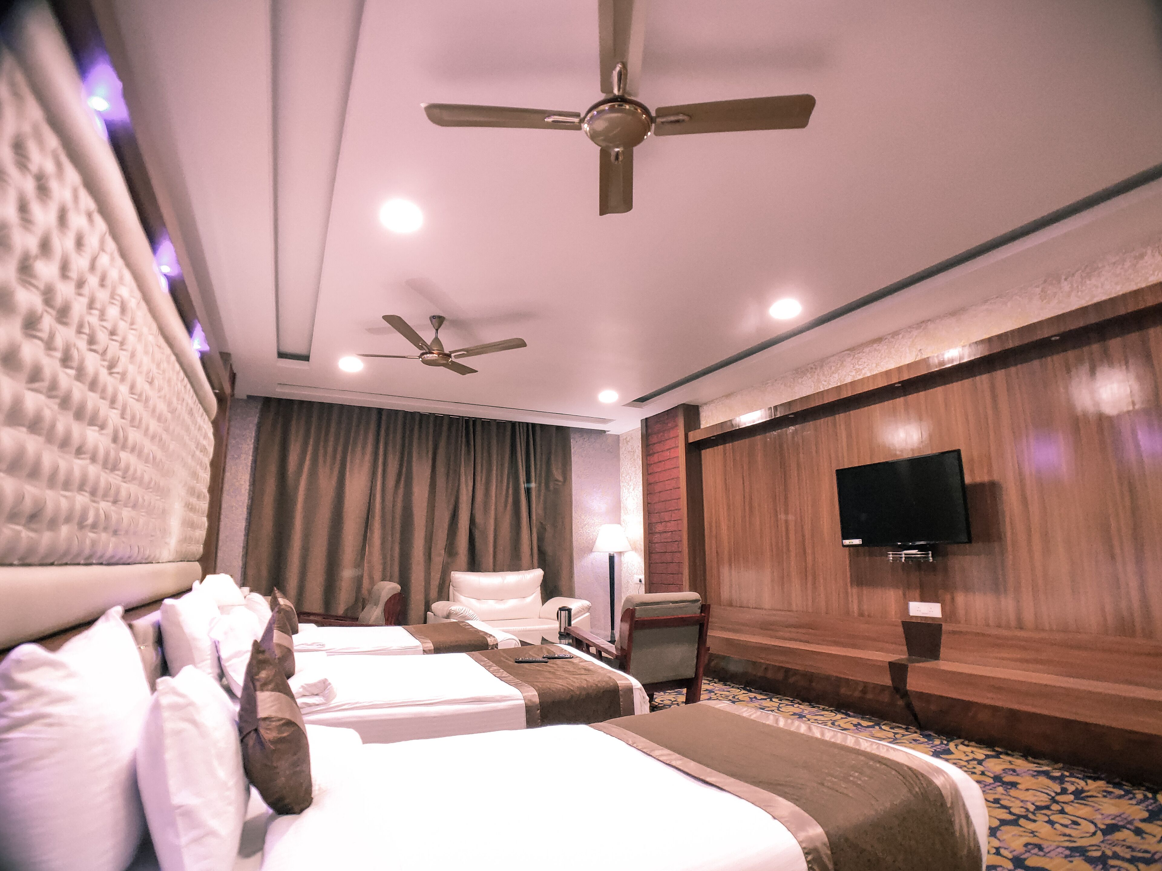 A hotel room with two beds, a ceiling fan, a TV mounted on the wall, and a carpeted floor.