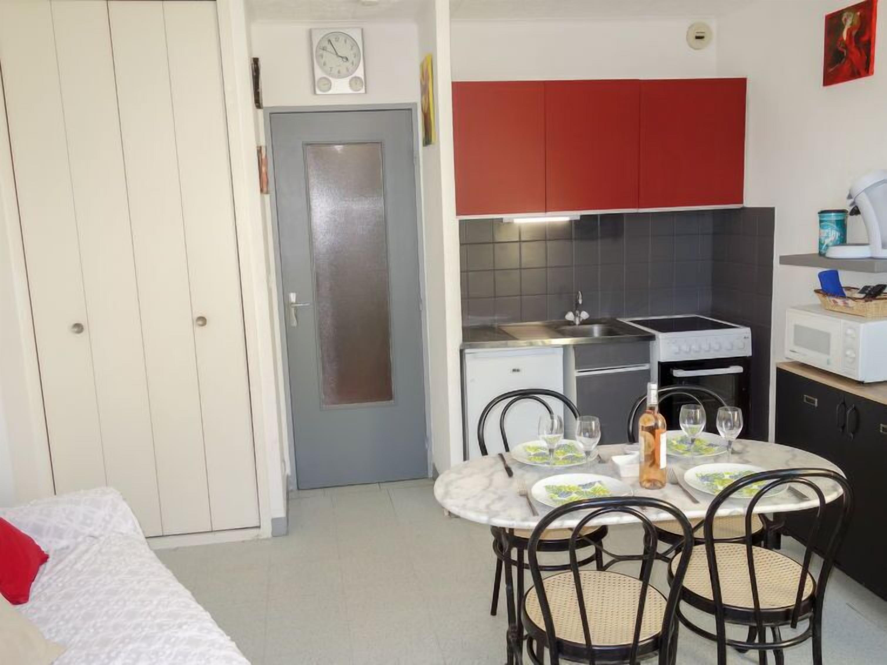 A compact kitchen with a stove, microwave, and sink. There is a dining table with chairs and a clock on the wall.
