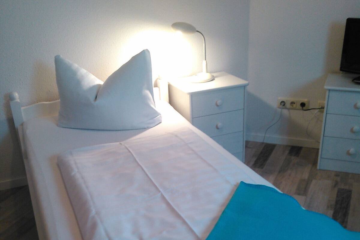 Un lit bien fait, avec du linge de lit blanc et une couverture bleue, une lampe de chevet et une petite table de chevet dotée de tiroirs.