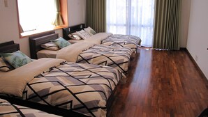 A hotel room with four beds aligned in a row, wooden flooring, and large windows with green curtains. - Hibisu yabu (Nago)