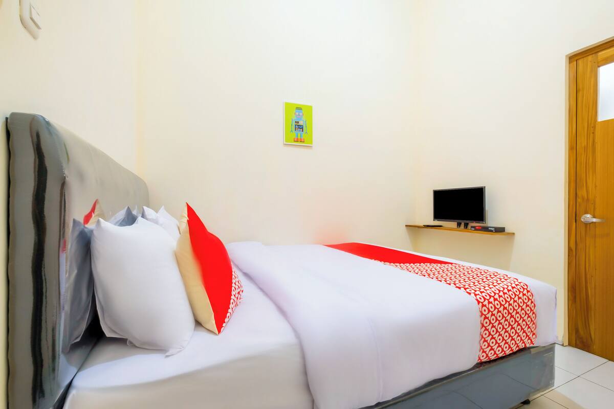 A bed with white and red bedding, a grey headboard, and a small TV on a wooden shelf above it.