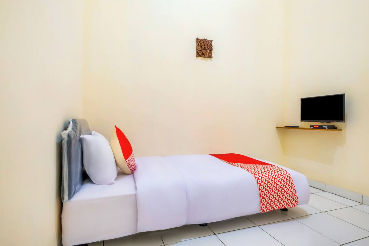 A single-bed room with a white mattress, red and white patterned blanket, and a television on a shelf above the bed.