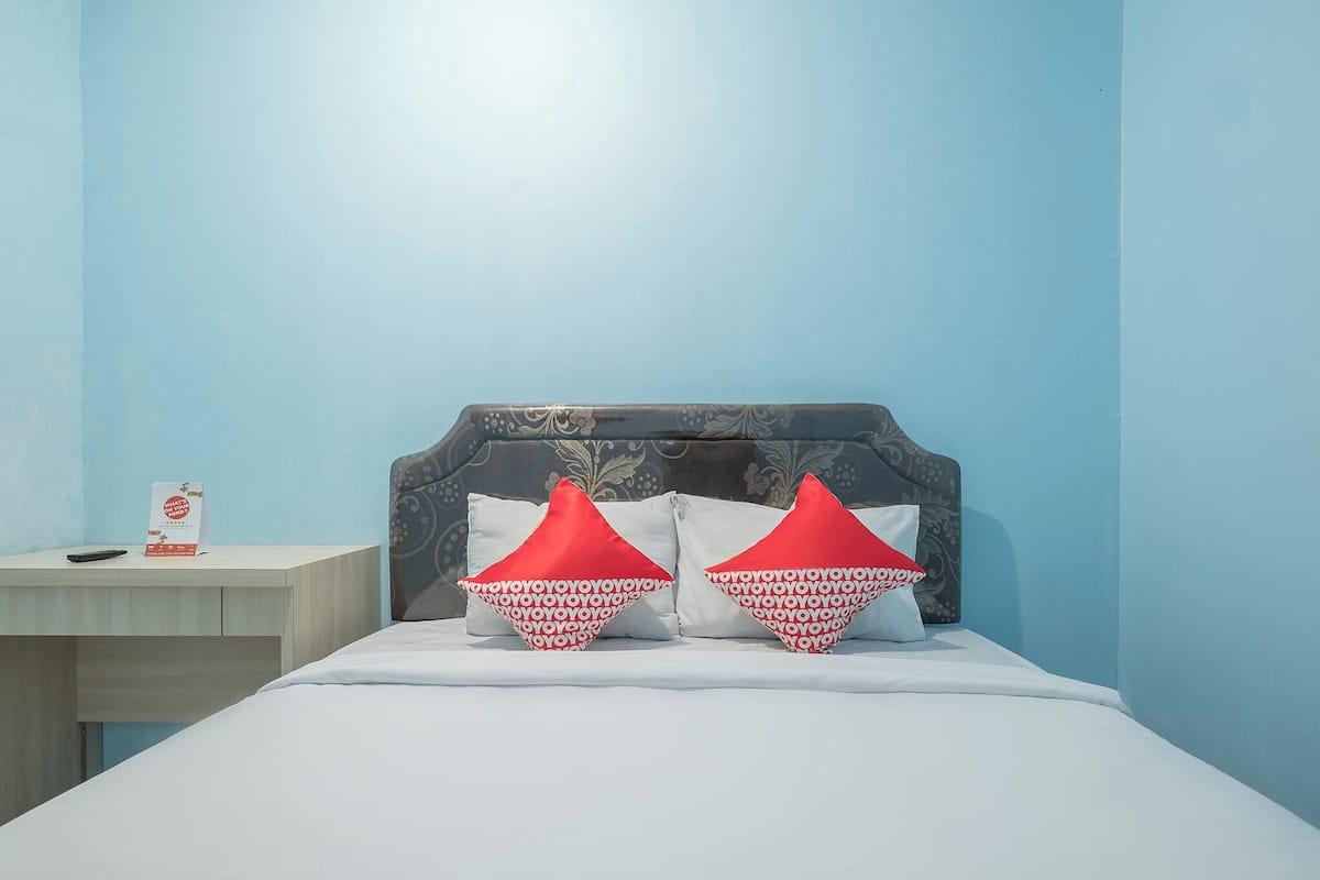 Un lit avec un drap blanc, deux oreillers rouges, une table de chevet avec une télécommande, et un mur orné d’un motif floral.
