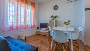 Una sala de estar moderna con un sofá azul, una mesa de comedor con sillas blancas, un televisor sobre un mueble de madera y una planta en maceta.