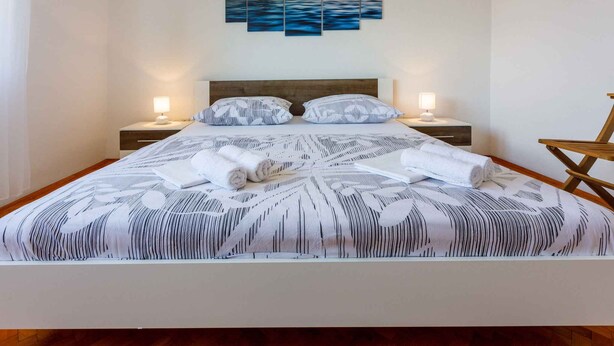 Una cama bien hecha con un edredón estampado, dos lámparas de noche y un cuadro de atardecer colgado en la pared.