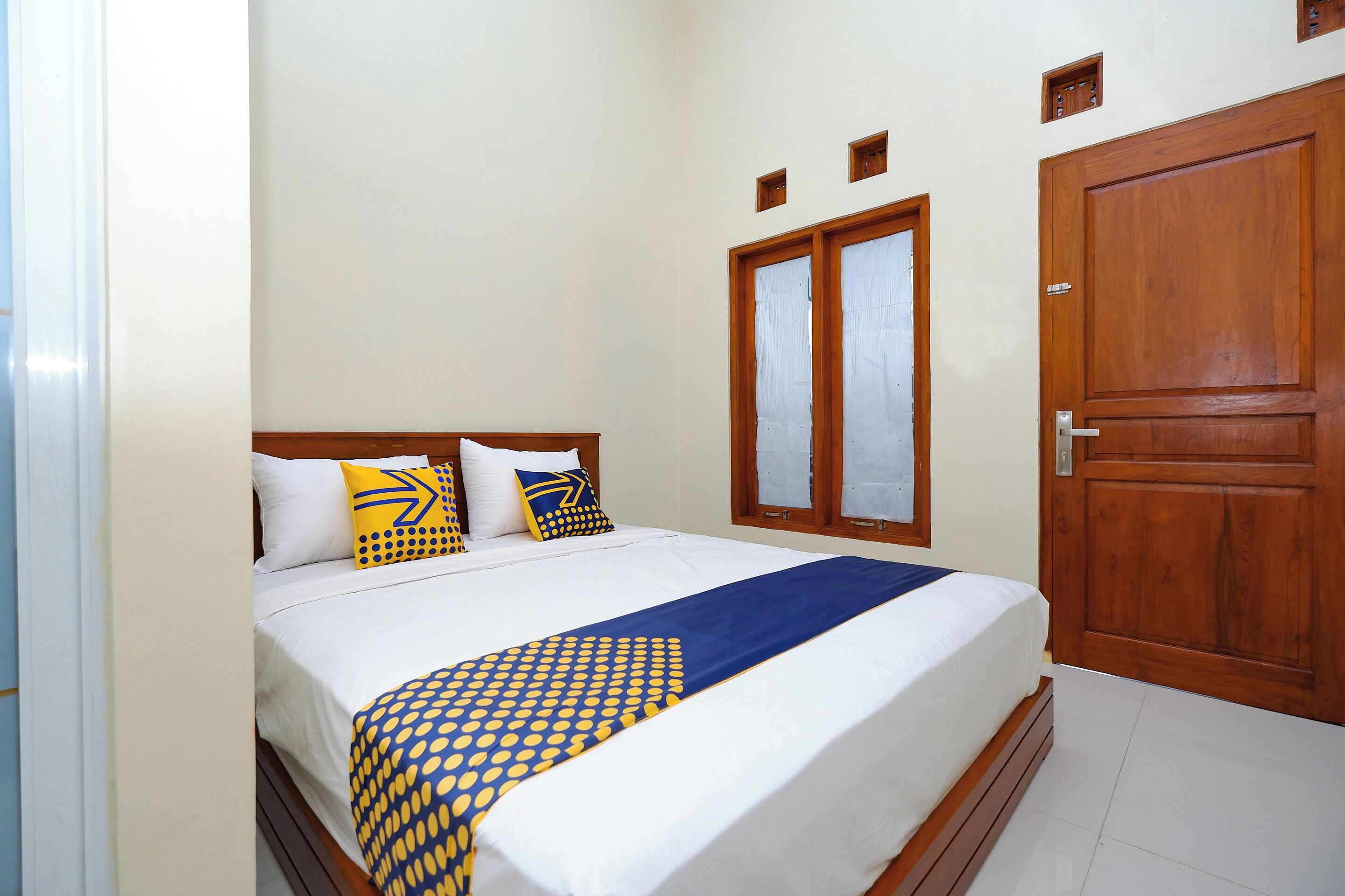 Una cama bien hecha con una colcha azul y amarilla estampada, un cabecero de madera y una puerta de madera con un ventanuco.