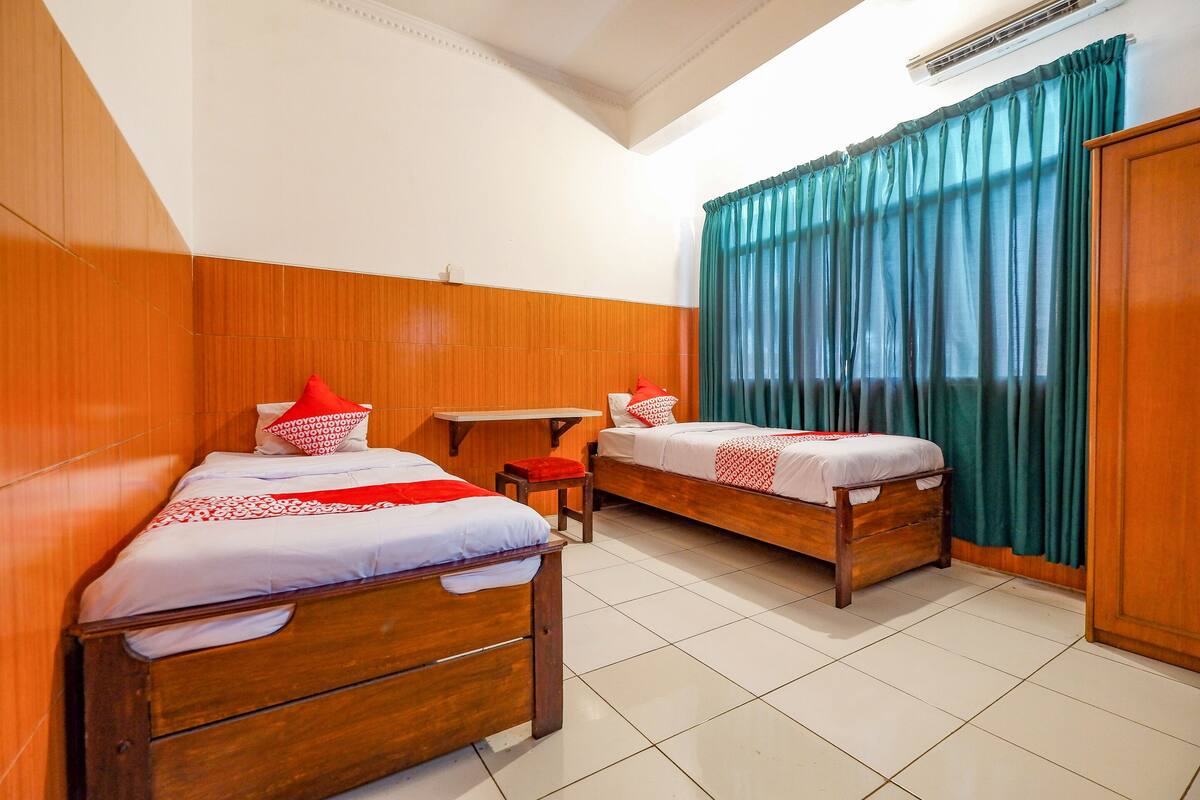 Two wooden beds with white bedding and red accents, a small table, and a window with teal curtains.