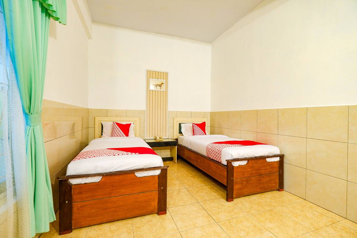 A twin room with two wooden beds, a small bedside table, a wall-mounted telephone, and a window with a curtain.