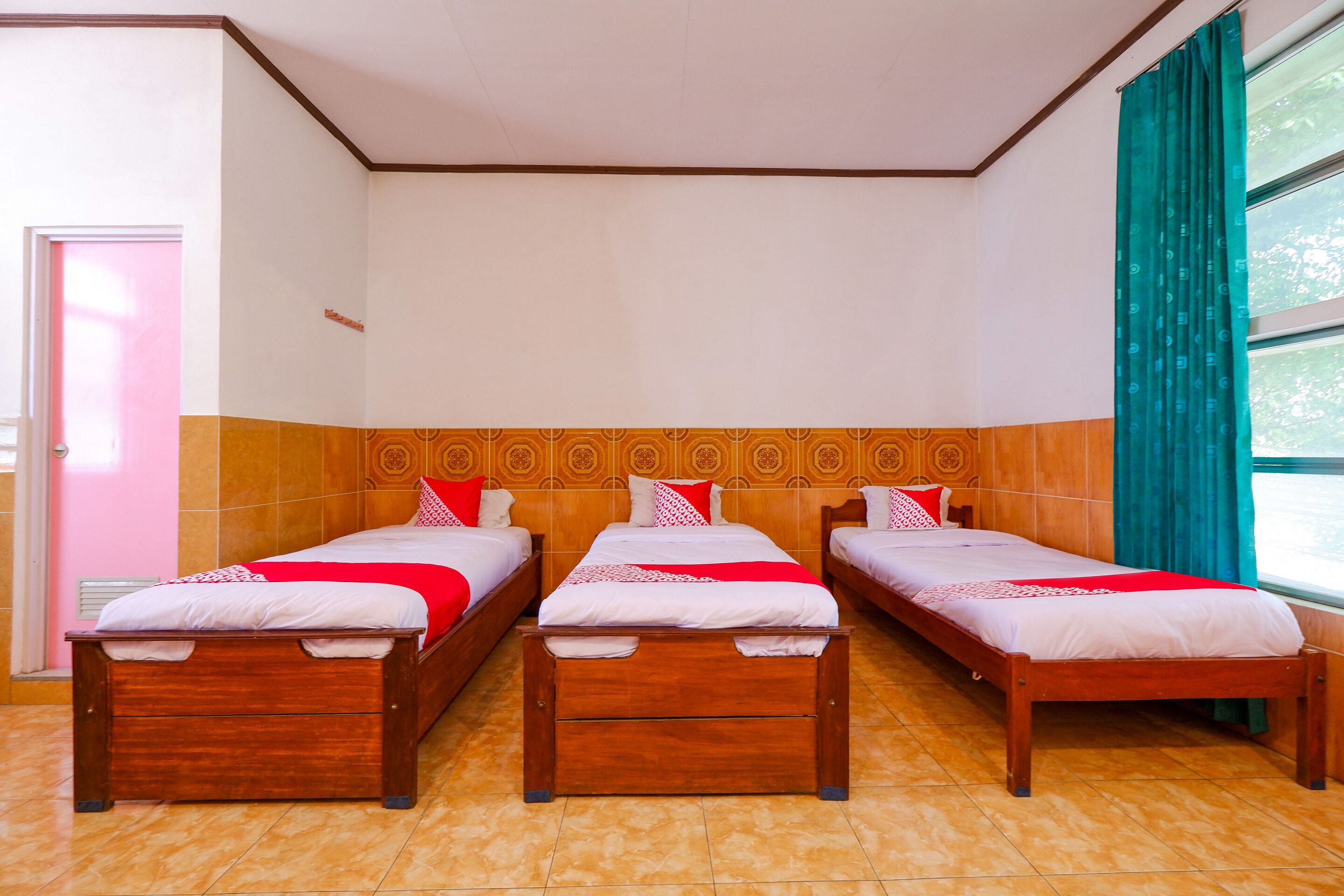Quarto de hotel com três camas de madeira, cada uma com colcha vermelha e branca, e parede laranja e branca com padrão.