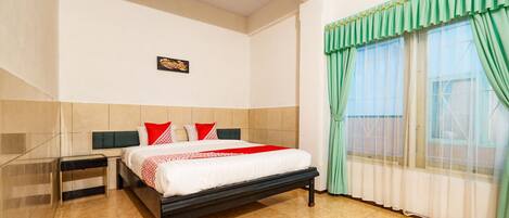 A hotel room with a bed, a nightstand, a window with green curtains, and a wall-mounted picture.