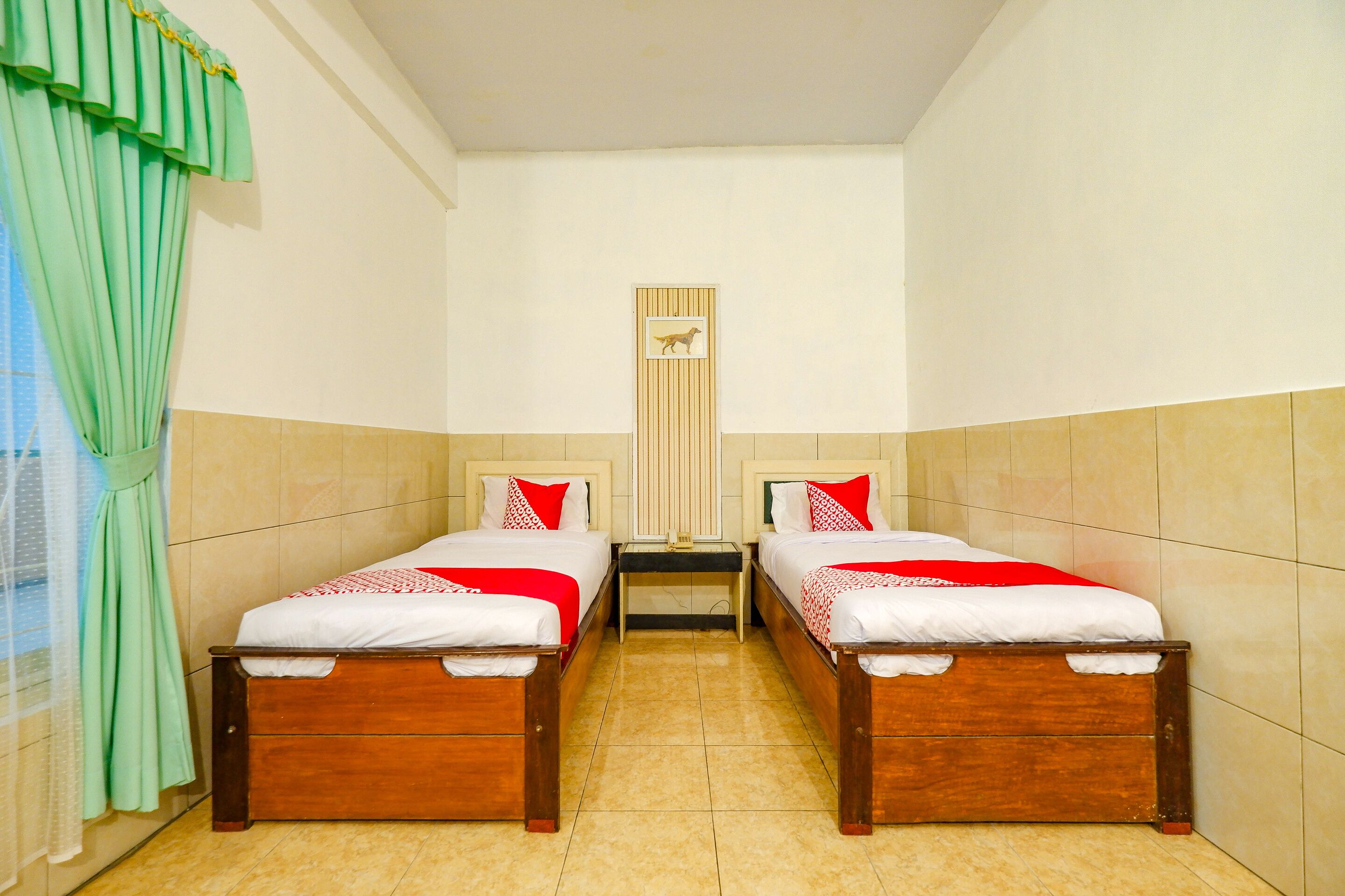 A room with two wooden beds, each with red and white bedding, a small table, and a window with green curtains.