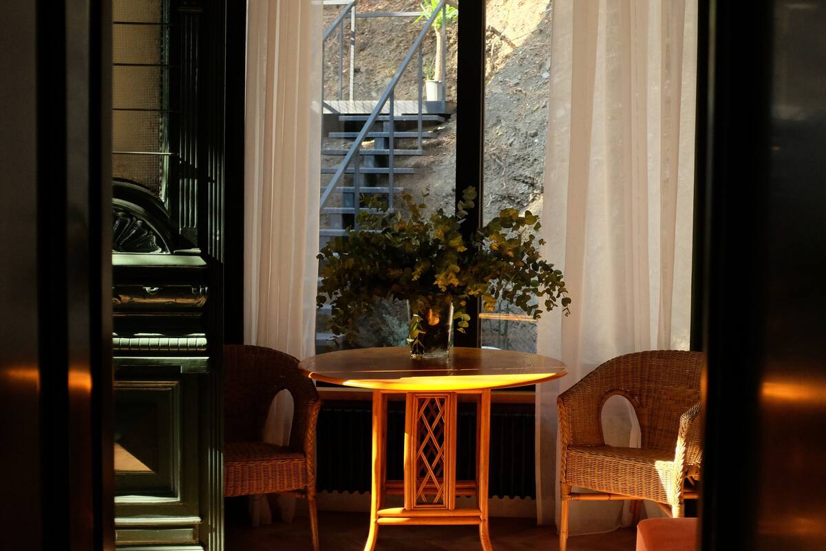 A room with a round table, two wicker chairs, a vase with green foliage, and a view of a staircase outside.