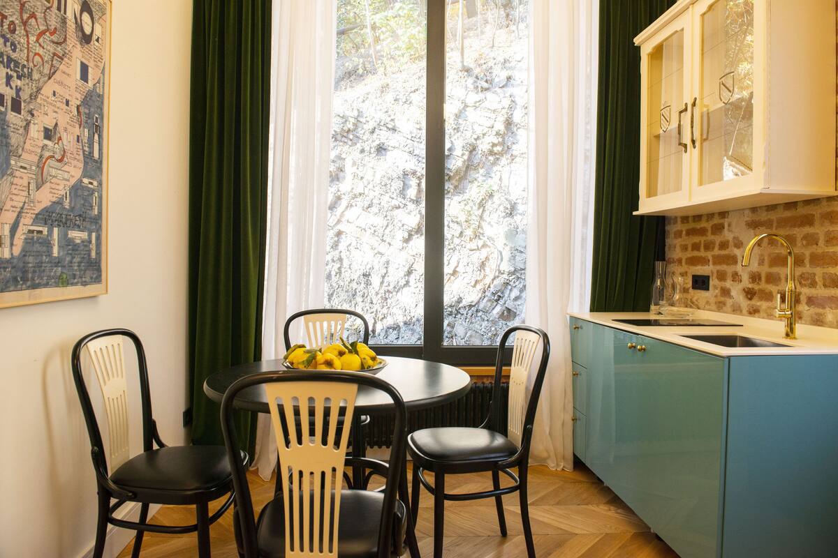 A dining area with a round table, four chairs, a bowl of fruit, a window with curtains, and a kitchen with blue cabinets and a sink.
