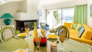 A bright living room with a yellow sofa, a fireplace, and a dining area with a glass table set for a meal. - Vacation home Hélios by Interhome (Port Camargue)