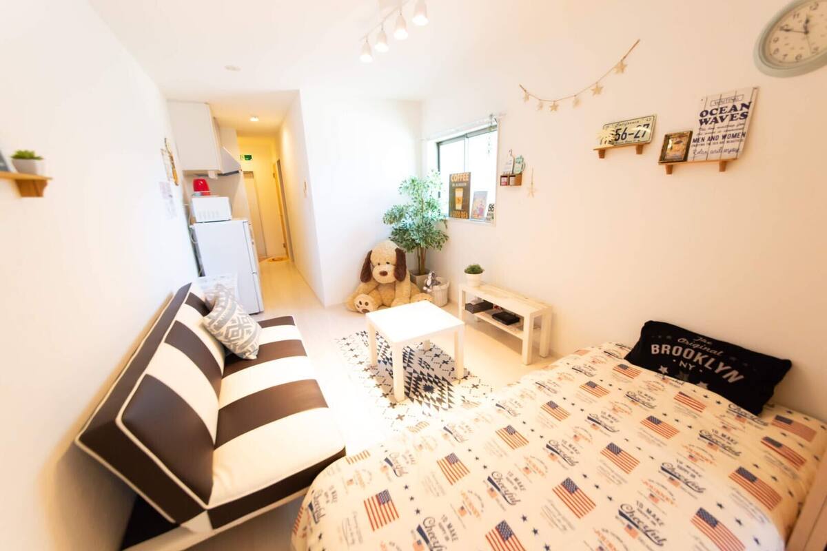 A cozy room with a striped sofa, a bed with a Brooklyn-themed bedspread, a small table, a potted plant, and a wall clock.