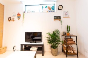 Una sala de estar con un televisor de pantalla plana sobre un mueble bajo, una planta en maceta, un estante con libros y un reloj, y un letrero en la pared con una placa de matrícula.