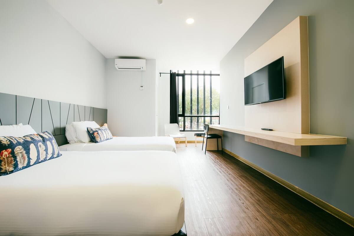 Ein Hotelzimmer mit zwei Betten, einem Schreibtisch, einem Stuhl, einem an der Wand befestigten Fernseher und einem Fenster mit Jalousien.