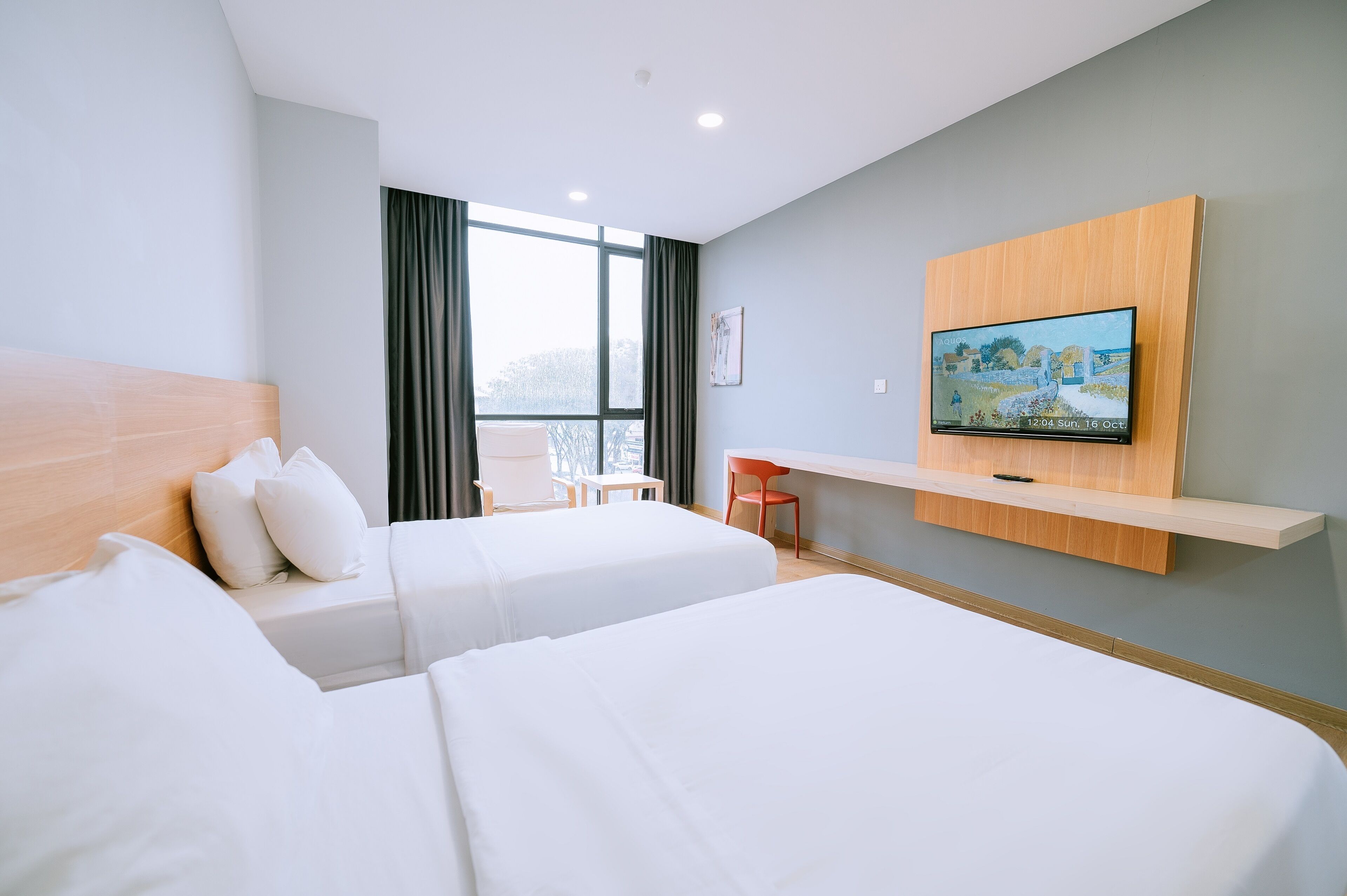 Habitación de hotel con dos camas, un televisor montado en un estante de madera, una silla y una ventana con cortinas.