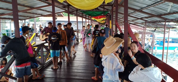 A covered walkway with people walking and standing, wooden railings, and a view of boats in the background. - Sipadan Inn 3 (Semporna)