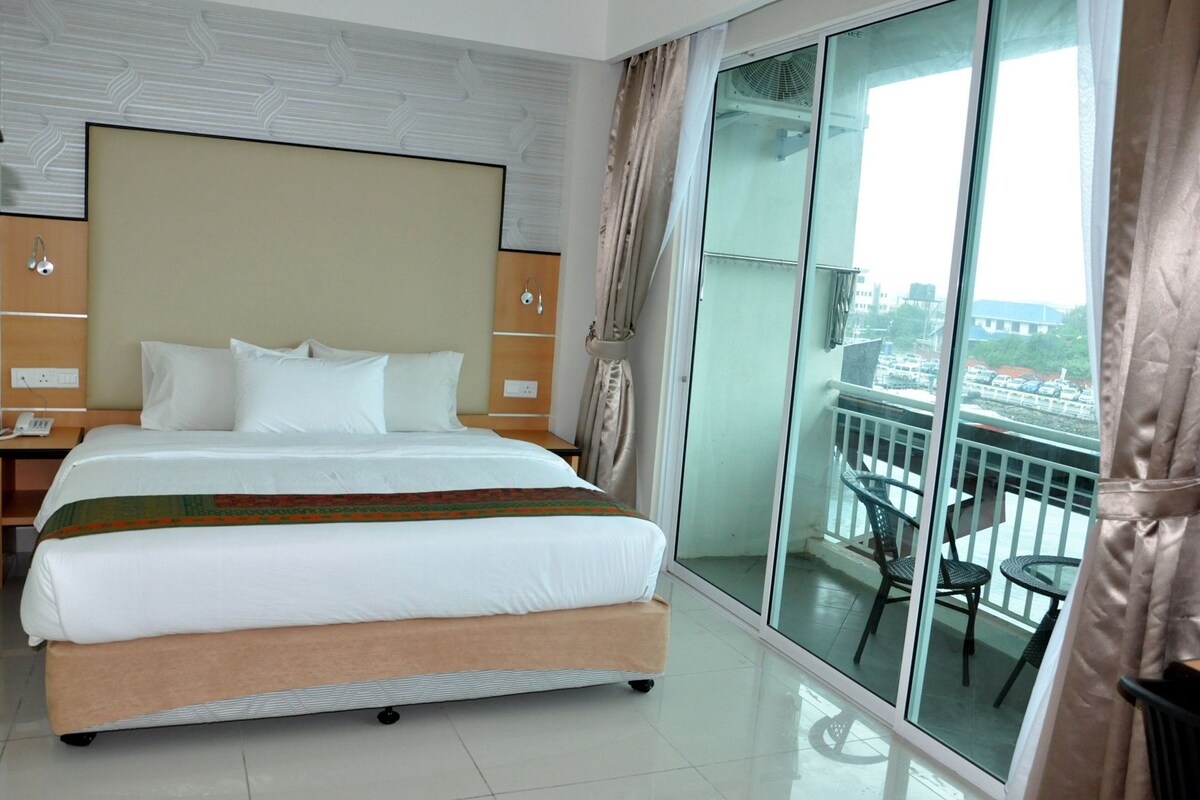 Habitación de hotel con una cama grande, una mesita de noche, un ventanal que da a un balcón con sillas y vistas a la ciudad.
