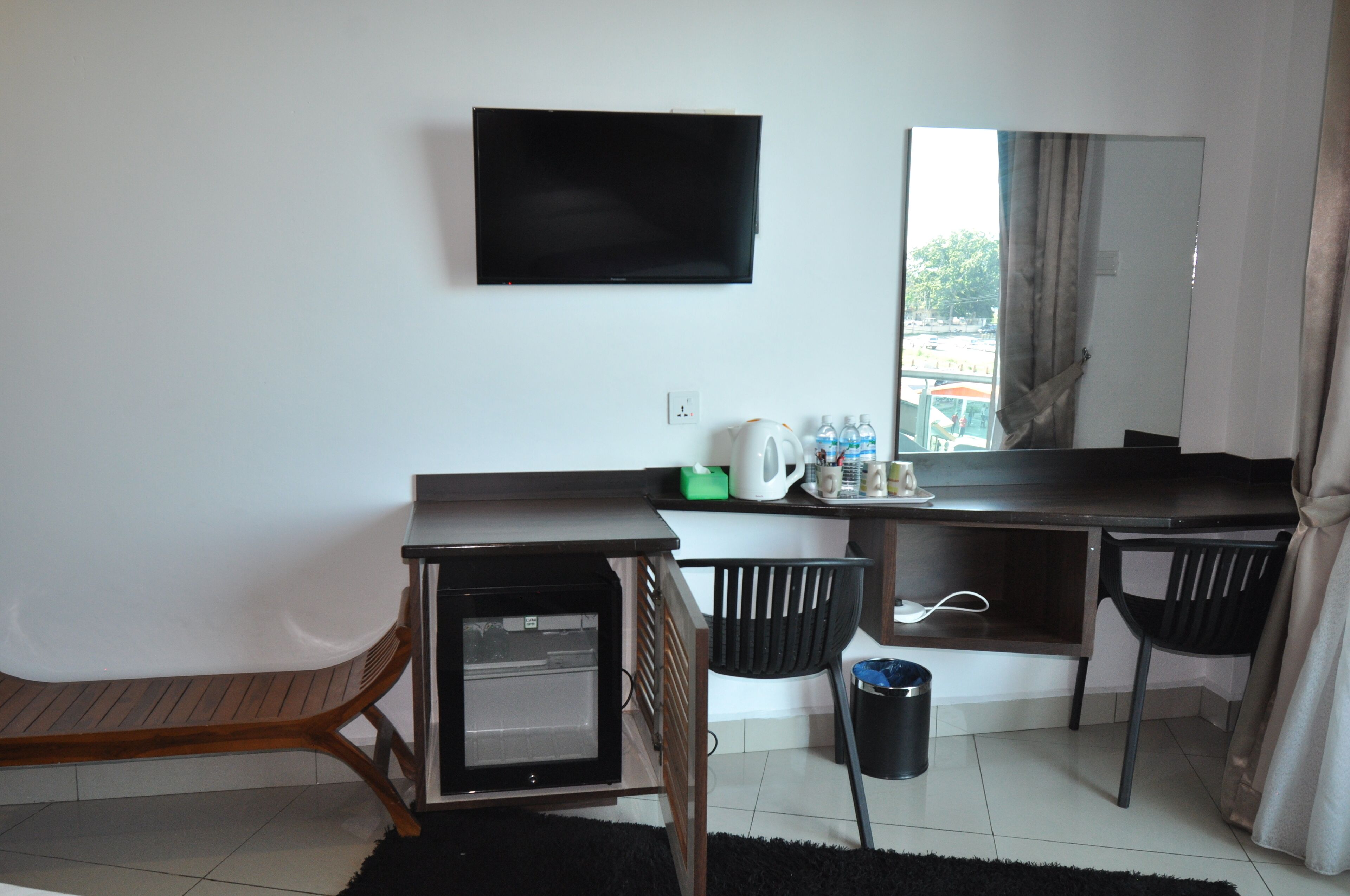 Ett kompakt hotellrum med en platt-TV som är monterad på väggen, ett litet skrivbord med en vattenkokare, koppar och en papperskorg.