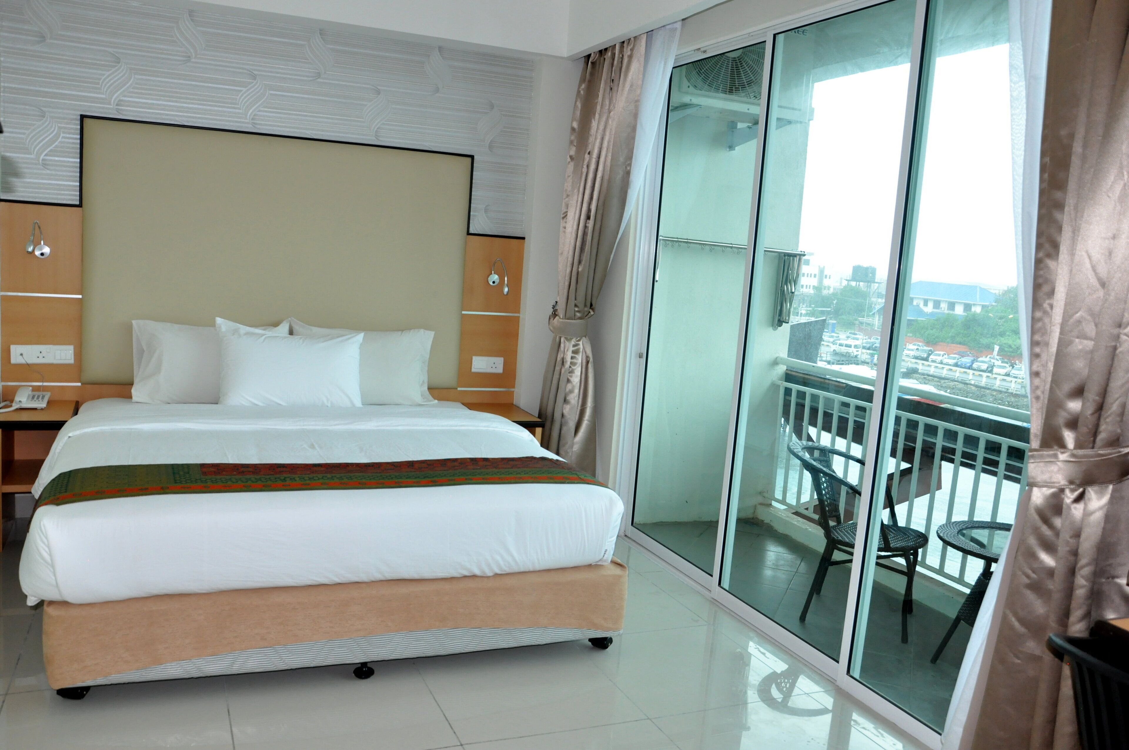 A hotel room with a large bed, a nightstand, a glass door leading to a balcony with chairs.