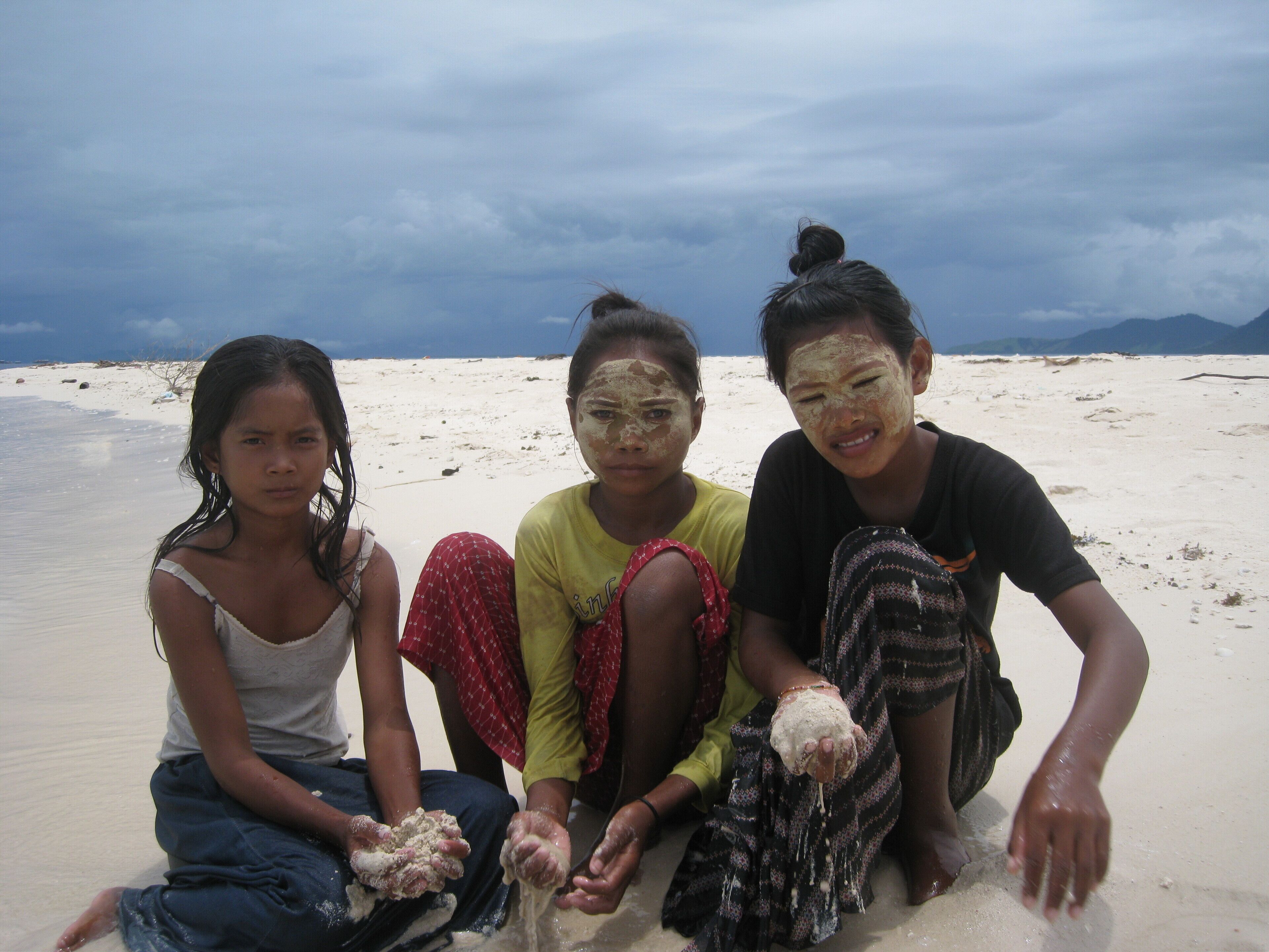Three individuals sitting on a sandy beach with a cloudy sky in the background.