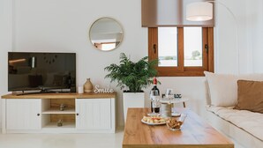 A living room with a TV, a wooden coffee table, a sofa, a plant, and a window with blinds. - Villa Oriens in Gennadi (Rhodes)