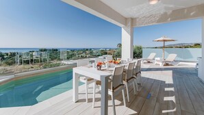 A poolside area with a table and chairs, a lounge chair, and a view of the sea. - Villa Oriens in Gennadi (Rhodes)