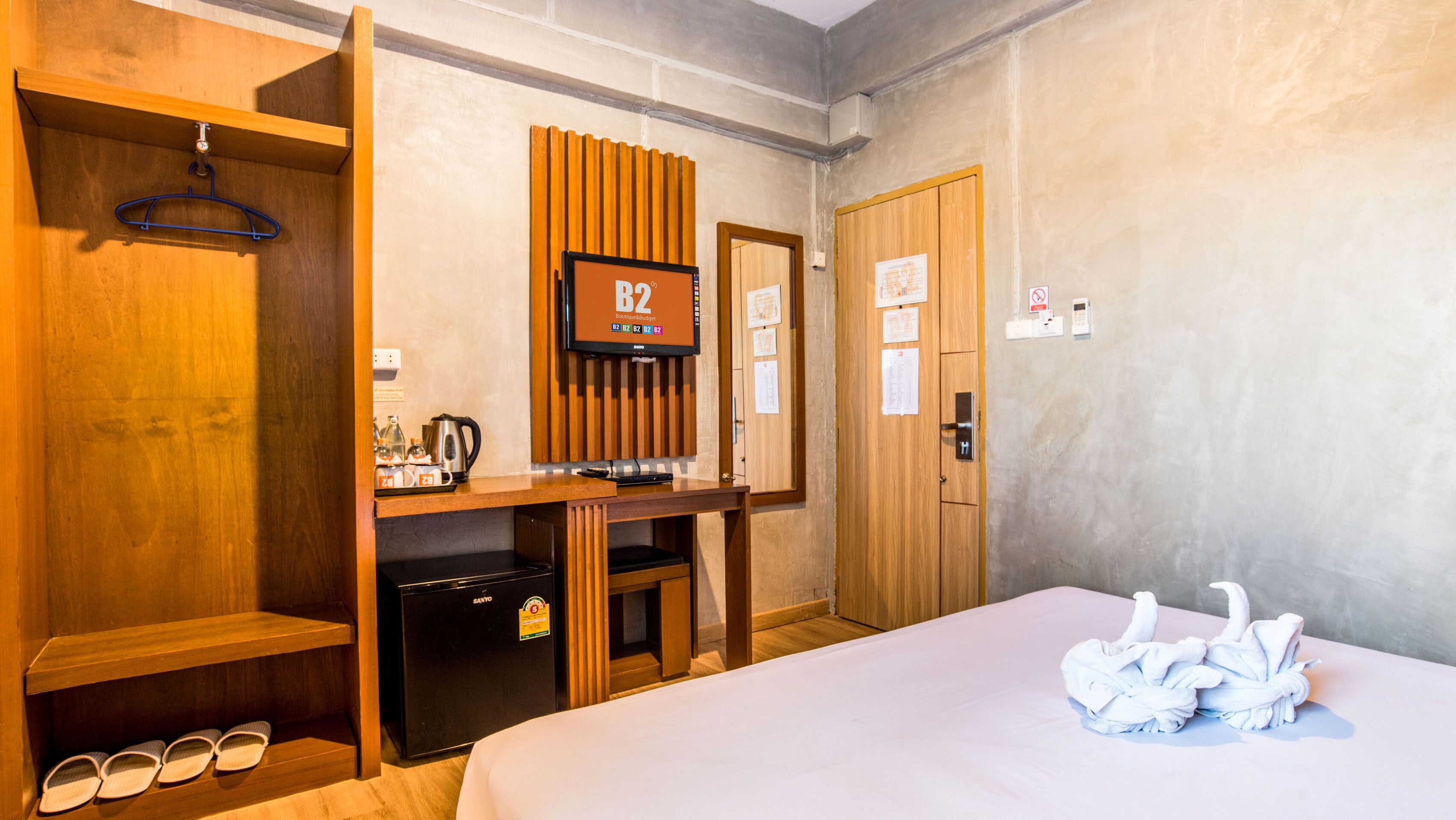 A hotel room with a bed, a desk with a computer, a minibar, a coat rack, and a door with a sign.