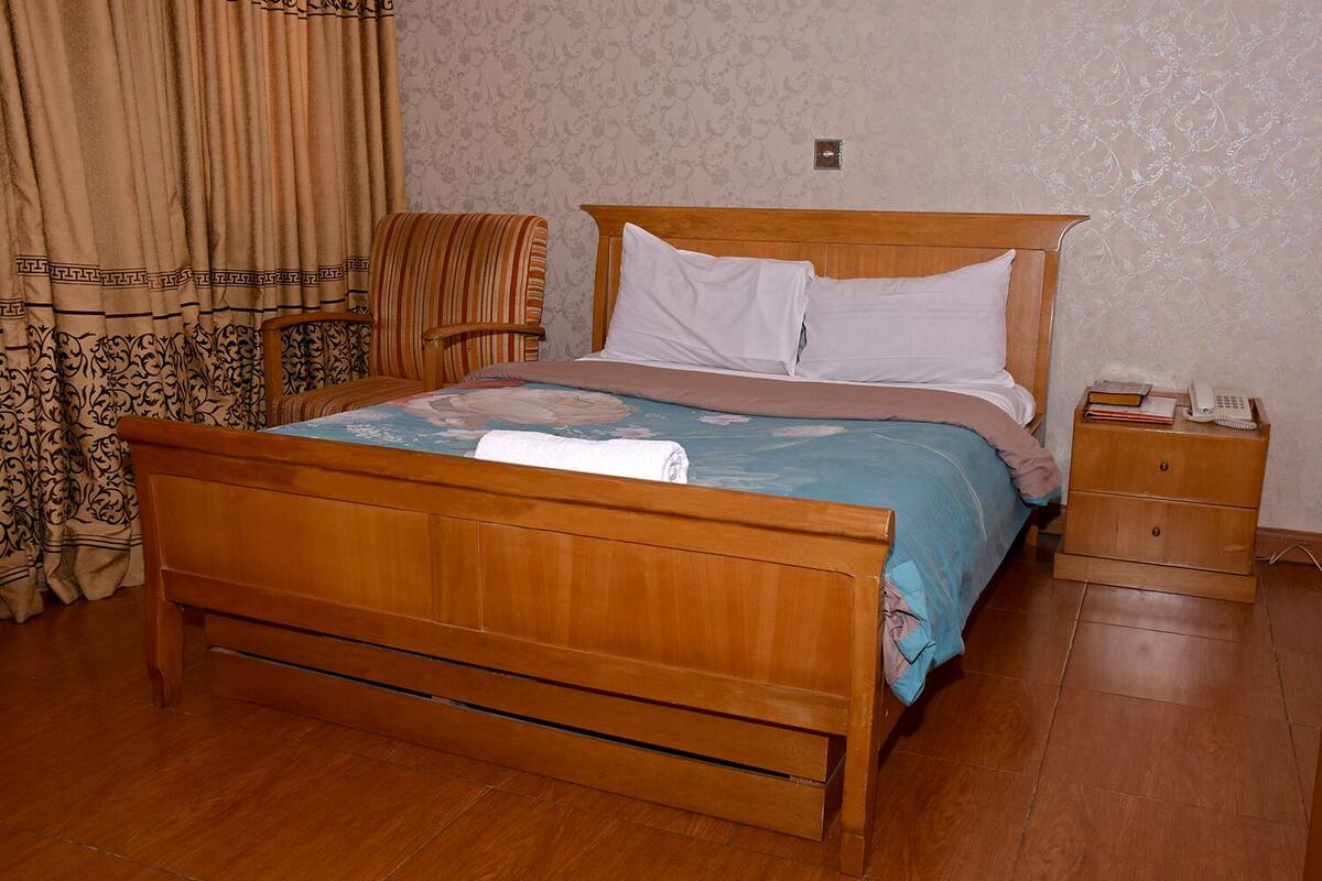 A wooden bed with a headboard, a bedside table with a phone and books, and a chair with a striped cushion.