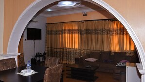 A living room with a sofa, armchairs, a coffee table, and a dining table with chairs. There is a TV mounted on the wall and a ceiling light fixture. - LaMaison Hotel (Ibadan)