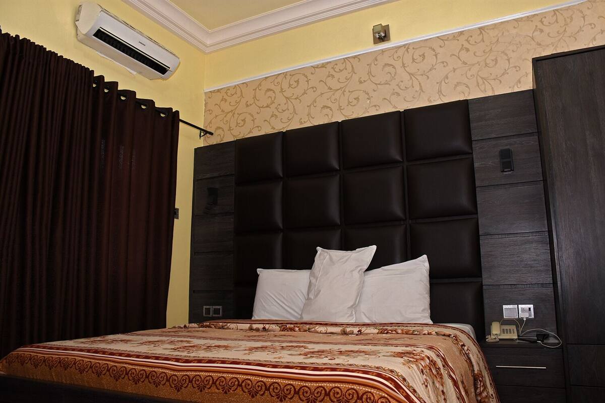 A bedroom with a dark headboard, a bed with white pillows, a patterned bedspread, and an air conditioning unit on the wall.