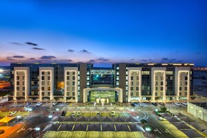 Um hotel moderno com fachada de vidro central, vários níveis de estacionamento e entrada bem iluminada.